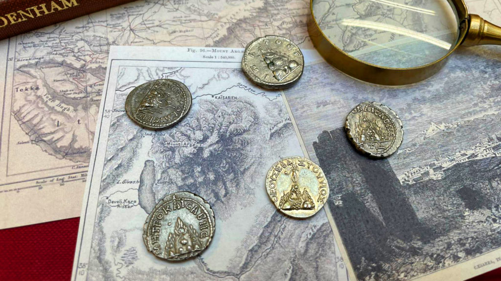 A few roman ancient coinage displayed on top of some ancient Turkish maps with a magnifying glass.